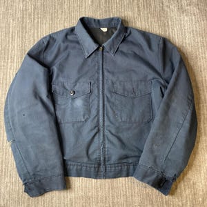 May include: A dark navy blue work jacket featuring a pointed collar, a full-length front zip, and two chest pockets with button-down flaps. The jacket has long sleeves with buttoned cuffs and shows signs of wear, including small tears on the sleeves.