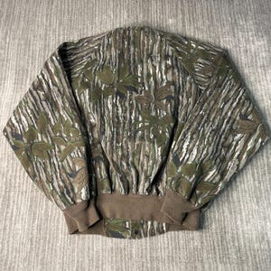 May include: A camouflage jacket with a brown ribbed collar and cuffs. The jacket features a pattern of vertical lines in shades of brown, green, and gray, with leaf-like shapes scattered across the fabric. The jacket has a button closure at the bottom.