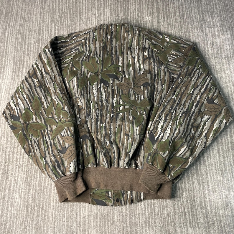 May include: A camouflage jacket with a brown ribbed collar and cuffs. The jacket features a pattern of vertical lines in shades of brown, green, and gray, with leaf-like shapes scattered across the fabric. The jacket has a button closure at the bottom.
