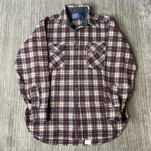 Puede incluir: Una camisa de manga larga con botones y un patrón de cuadros en tonos burdeos, crema y azul oscuro. La camisa incluye un forro de cuello acolchado en verde oscuro, dos bolsillos en el pecho con solapas abotonadas y cierre frontal de botones.