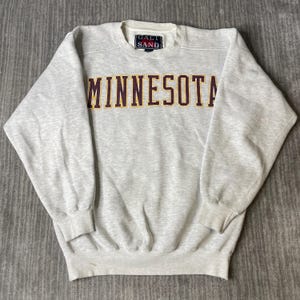 Pode incluir: Uma camisola de gola redonda em cinzento mesclado claro com a palavra MINNESOTA em letras grandes e arrojadas de cor bordeaux escuro com um contorno amarelo fino. A peça tem uma etiqueta da marca Galt Sand no interior da gola e mangas compridas.