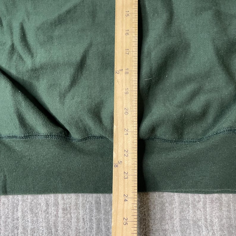 May include: Close-up of a dark green sweatshirt with a ribbed hem and cuffs. A wooden ruler is placed vertically along the right side, showing measurements in inches. The fabric appears to be a soft, knit material.