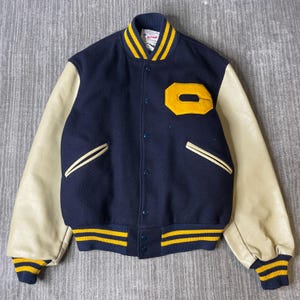 May include: A navy blue wool varsity jacket with cream-colored leather sleeves. The jacket features a yellow letter O patch on the chest, a snap-button front closure, and striped yellow and navy blue ribbed trim on the collar, cuffs, and hem.