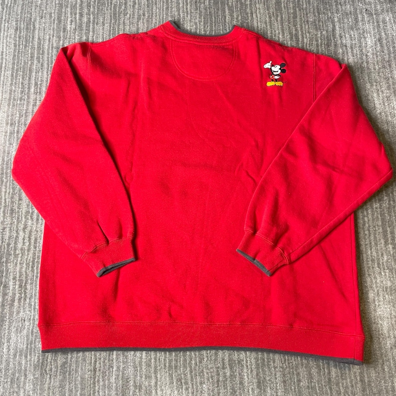 Puede incluir: Una sudadera roja de manga larga con un peque&ntilde;o gr&aacute;fico bordado de Mickey Mouse en la parte superior derecha del pecho. La prenda tiene cuello redondo, pu&ntilde;os acanalados y un dobladillo acanalado, todo acabado con un borde gris oscuro.