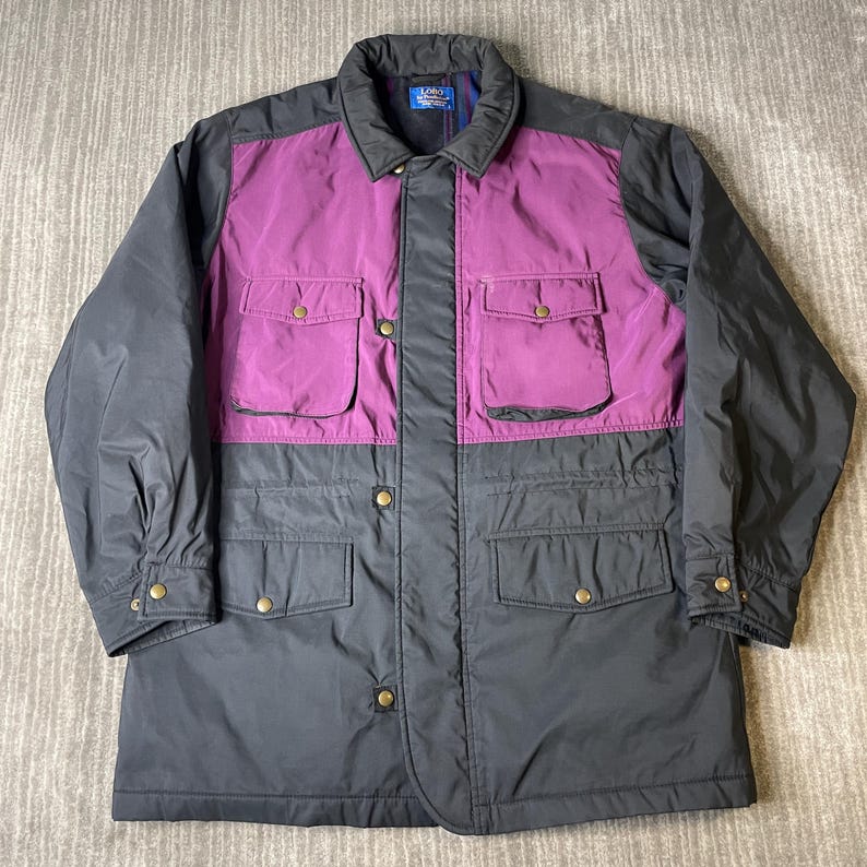 K&ouml;nnte beinhalten: Zweifarbige Jacke mit lila Oberbrust und Taschenbereich sowie einem dunkelgrauen unteren Bereich. Die Jacke hat einen Kragen, vier Vordertaschen und goldfarbene Knopfverschl&uuml;sse. Der Markenname "Zory" ist sichtbar.