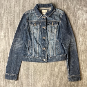 May include: A medium-wash blue denim jacket featuring a classic collar, long sleeves with buttoned cuffs, and a front button closure. The garment includes two chest pockets with buttoned flaps and two vertical side pockets, with a Mudd brand label.