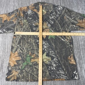 May include: A camouflage-patterned t-shirt with brown, green, and gray tones. The shirt features a realistic forest design with leaves and tree bark. A wooden ruler is placed across the shirt for measurement.