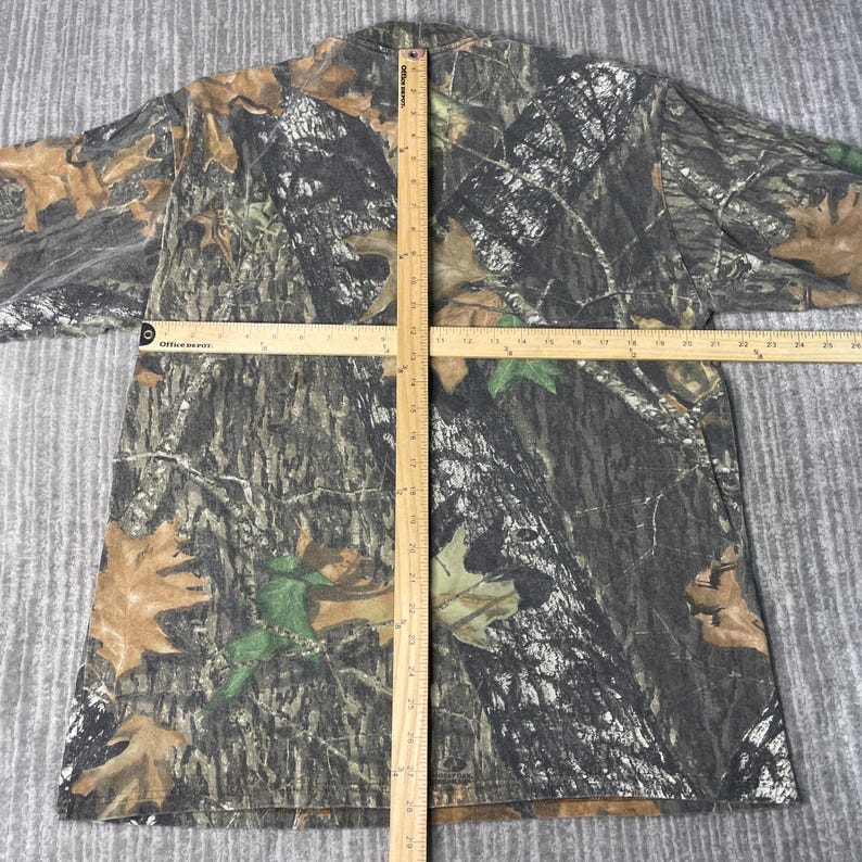 May include: A camouflage-patterned t-shirt with brown, green, and gray tones. The shirt features a realistic forest design with leaves and tree bark. A wooden ruler is placed across the shirt for measurement.