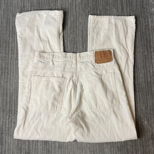 May include: Off-white corduroy pants featuring a straight-leg cut, a high-rise waist with belt loops, two back patch pockets, and a tan leather-like brand patch sewn onto the rear waistband. The fabric has a distinct vertical ribbed texture.