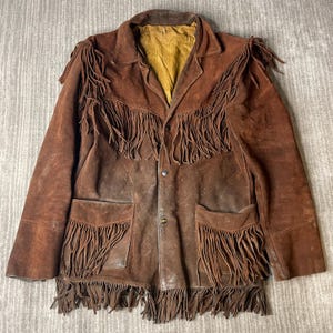 May include: A brown suede jacket featuring long fringe detailing across the chest, shoulders, and hem. The jacket includes two front pockets with matching fringe, a collared neckline, and a mustard yellow interior lining with three front buttons.