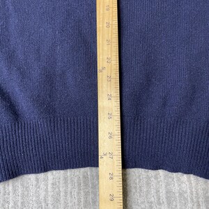 Puede incluir: Un su&eacute;ter de punto azul marino con dobladillo acanalado, colocado junto a una regla de madera que muestra medidas de 48 cm a 74 cm, proporcionando una referencia visual clara de la longitud de la parte inferior y el dobladillo.