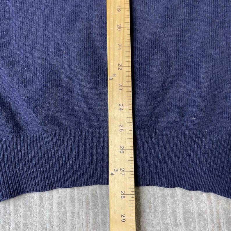 Puede incluir: Un su&eacute;ter de punto azul marino con dobladillo acanalado, colocado junto a una regla de madera que muestra medidas de 48 cm a 74 cm, proporcionando una referencia visual clara de la longitud de la parte inferior y el dobladillo.