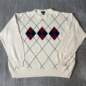 May include: Cream-colored sweater with a classic argyle pattern. The sweater features a diamond design in navy blue, burgundy, and cream. The sweater has a crew neck and long sleeves.
