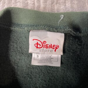 May include: Close-up of a green sweatshirt collar and a Disney Store label. The label features the Disney logo in red, the word "store", and text indicating the size is S and the decoration is exclusive of Brunei.