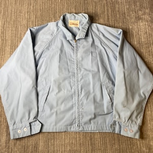 May include: A light blue zip-up jacket featuring a pointed collar, long sleeves with buttoned cuffs, and two front pockets. The interior neck label displays the text King Louie. The garment is constructed with raglan sleeves and a central zipper.