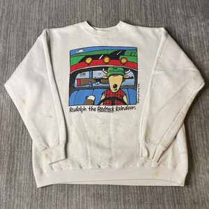 May include: Cream-coloured sweatshirt with a cartoon image of a reindeer driving a car, with the text "Rudolph the Redneck Reindeer." The reindeer is wearing a plaid shirt and a green hat. A rifle is also visible.