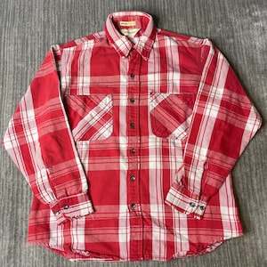 May include: A long-sleeve button-down shirt featuring a red and white plaid pattern. The shirt includes a pointed collar, two chest pockets with button closures, and buttoned cuffs. The front placket has a series of dark buttons down the center.