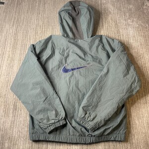 May include: A sage green hooded windbreaker jacket featuring a dark blue Nike swoosh logo centered on the back. The jacket has long sleeves with elastic cuffs, a matching elastic hem, and a contrasting dark gray panel running up the hood.