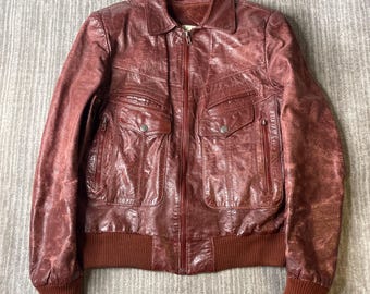 May include: A dark red leather jacket featuring a full-length front zip, a standard collar, and long sleeves with ribbed knit cuffs. The jacket includes two chest pockets with snap-button flaps, two vertical zipped pockets, and a ribbed hem.