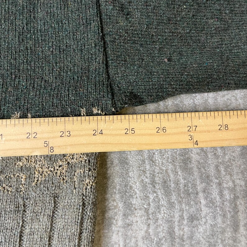 K&ouml;nnte beinhalten: Nahaufnahme eines dunkelgr&uuml;nen Stoffes mit einem Holzlineal. Das Lineal zeigt Messungen von 53 cm bis 71 cm. Der Stoff hat eine strukturierte Optik.