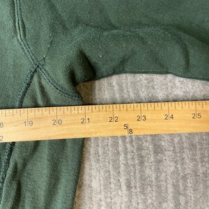 May include: Close-up of a dark green sweatshirt with a wooden ruler measuring the fabric. The ruler shows measurements from 48.3 cm to 63.5 cm. The fabric has visible stitching and a slightly worn texture.