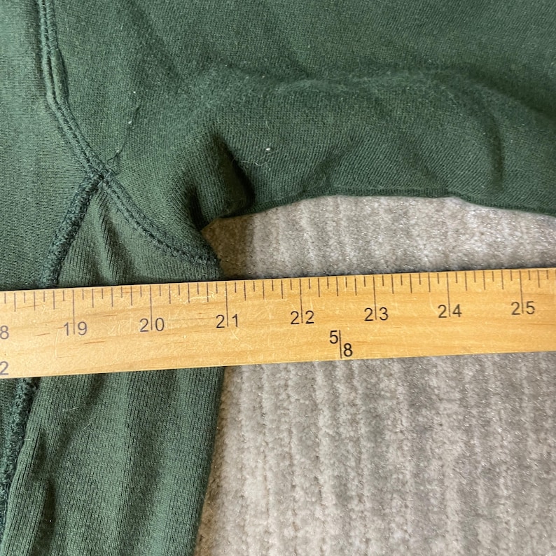 May include: Close-up of a dark green sweatshirt with a wooden ruler measuring the fabric. The ruler shows measurements from 48.3 cm to 63.5 cm. The fabric has visible stitching and a slightly worn texture.