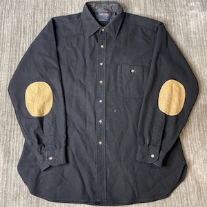 May include: A long-sleeve black button-down shirt featuring a pointed collar, a single chest pocket, and tan-colored oval elbow patches. The shirt has a front button placket with matching buttons and a curved hemline at the bottom of the garment.