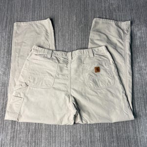 May include: Khaki-colored work pants with multiple pockets and a brown logo patch on the back. The pants are made of a durable material and have a relaxed fit. The pants are laid flat on a gray surface.