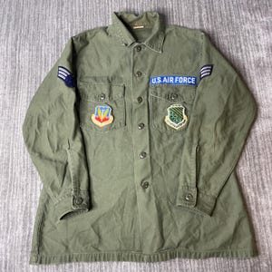May include: An olive green long-sleeve button-up shirt featuring a blue U.S. AIR FORCE patch, two chest pockets with flaps, and various embroidered military-style patches on the chest and sleeves, including a shield-shaped emblem on the right.