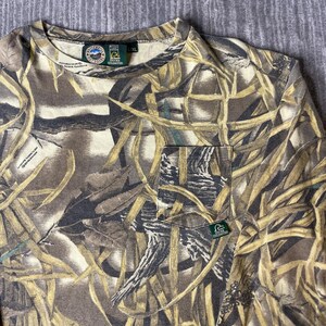 K&ouml;nnte beinhalten: Ein lang&auml;rmeliges T-Shirt mit einem Tarnmuster aus braunen, beigen und cremefarbenen Schilfhalmen und Bl&auml;ttern. Das Shirt verf&uuml;gt &uuml;ber eine Brusttasche mit einem kleinen gr&uuml;nen Ducks Unlimited-Etikett sowie einen Rundhalsausschnitt mit Innenetiketten.