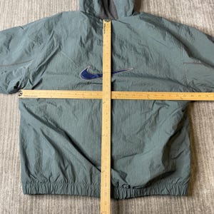 May include: A sage green Nike hooded windbreaker jacket with a dark blue swoosh logo on the chest. Two wooden rulers are placed over the garment, showing a width of approximately 26 inches and a vertical length of approximately 28 inches.