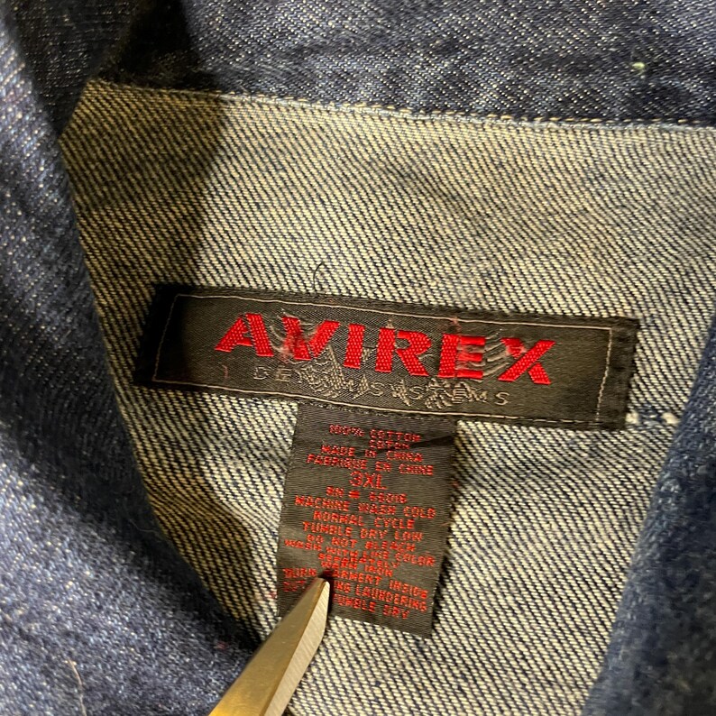 Pode incluir: Uma jaqueta jeans azul escuro com uma etiqueta interna preta com o texto vermelho AVIREX e o subt&iacute;tulo DENIM SYSTEMS. Uma etiqueta de cuidados preta abaixo exibe 100% COTTON, MADE IN CHINA, 3XL e v&aacute;rias instru&ccedil;&otilde;es de lavagem.