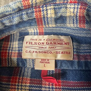 May include: Close-up of a blue, red, and white plaid flannel shirt. The shirt's label reads "FILSON GARMENT SINCE 1897 C.C.FILSONCO.-SEATTLE" and "MADE IN PORTUGAL L". The shirt is a size large.