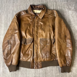 May include: A brown leather bomber jacket featuring a classic collar, a front zipper closure, two large flap pockets on the front, and dark brown ribbed knit cuffs and hem. The leather has a distressed finish with visible natural variations.