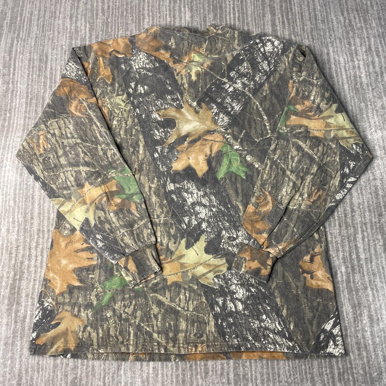 May include: Long-sleeved camouflage shirt featuring a pattern of brown, green, and tan leaves and branches. The shirt has a crew neck and long sleeves, suitable for outdoor activities or casual wear. The fabric appears to be a blend, offering comfort and durability.