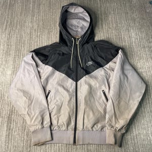 May include: A black, gray, and light gray Nike windbreaker jacket with a hood. The jacket has a chevron design across the chest and the Nike logo is visible on the upper left chest. The jacket has a full zipper and two side pockets.