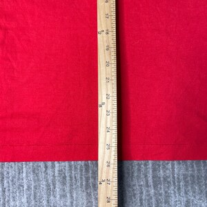 May include: A close-up of red fabric with a wooden ruler placed vertically. The ruler shows measurements from 16 to 28 inches. The fabric is a solid, vibrant red colour.
