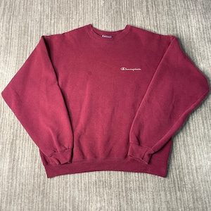 May include: A maroon Champion sweatshirt with a crew neck and long sleeves. The Champion logo is embroidered in white on the upper left chest. The sweatshirt is made of a soft, comfortable material.
