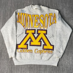 May include: A gray crewneck sweatshirt featuring the word "MINNESOTA" in yellow block letters above a large yellow and maroon "M" logo. The words "Golden Gophers" are below the logo. The sweatshirt has signatures in blue ink.