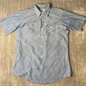 Puede incluir: Una camisa de manga corta de color azul claro con cuello en punta, dos bolsillos en el pecho con cierre de botón y una tapeta frontal con botones. La etiqueta de la marca es visible en el cuello interior con el texto BIG MAC.