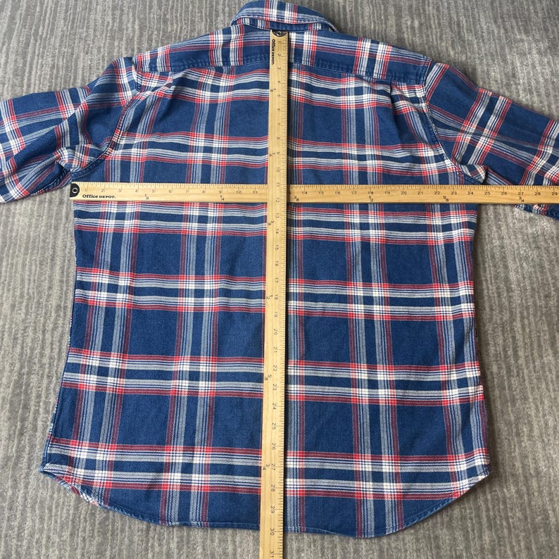 May include: A blue, red, and white plaid flannel shirt. The shirt is laid flat with a ruler across the chest and down the center. The shirt has a button-down collar and long sleeves. The fabric is a soft, woven material.