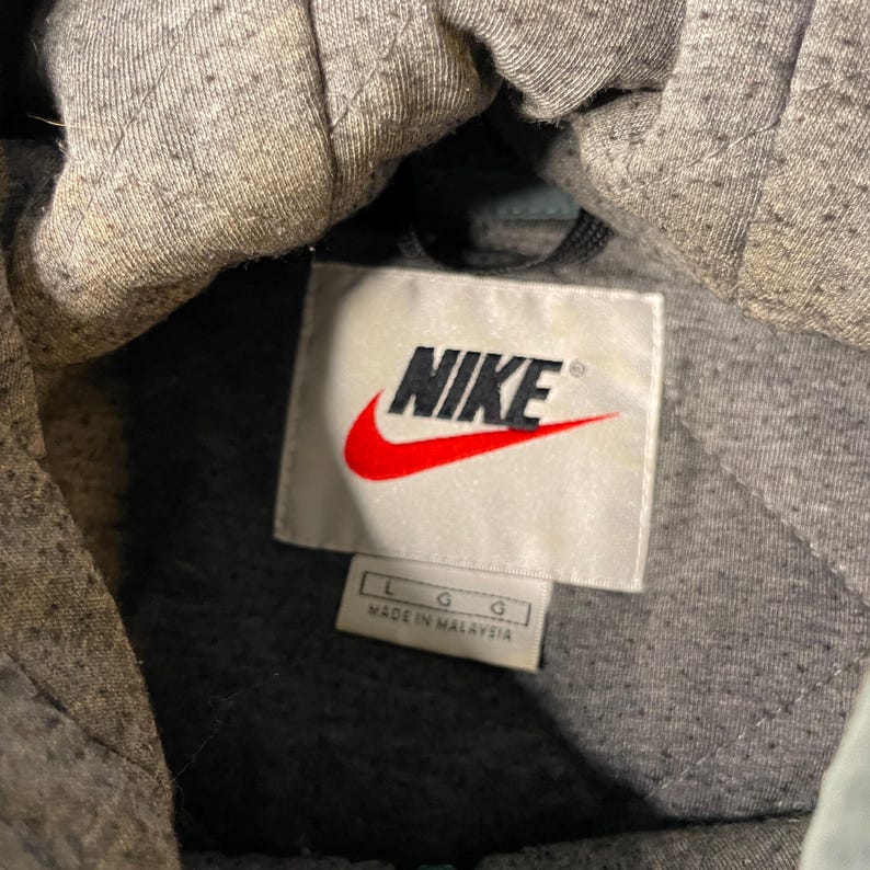 May include: A grey hooded sweatshirt with a quilted interior lining. A white rectangular tag is sewn into the neck area featuring the black Nike logo with a red swoosh. A smaller white tag below displays the size L and the text MADE IN MALAYSIA.