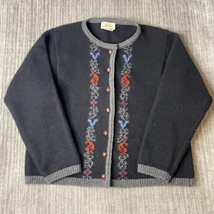 May include: A black long-sleeve cardigan featuring a round neckline and a front button closure. The garment is accented with two vertical panels of gray, blue, orange, and purple floral patterns. It has gray ribbed trim on the cuffs, hem, and collar.