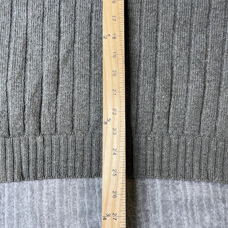 K&ouml;nnte beinhalten: Nahaufnahme eines grauen Rippstrickstoffs mit einem Holzlineal an der rechten Seite. Das Lineal zeigt Messungen in Zoll, von 16 bis 27 Zoll (ca. 40,6 bis 68,6 cm). Der Stoff hat vertikale Rippen.