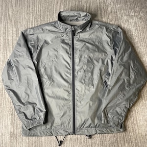 May include: A silver-grey windbreaker jacket featuring a high collar, long sleeves with elastic cuffs, and a full-length black front zipper. The lightweight fabric has a subtle grid pattern, and the hem includes an adjustable drawstring cord.