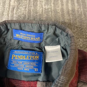 Op de afbeelding: Detail van de binnenkraag van een Pendleton Western Wear shirt. Het shirt heeft een rood en grijs ruitpatroon. Blauwe labels tonen "High Grade Western Wear" en "Pendleton 100% Pure Virgin Wool". Gemaakt in de VS.