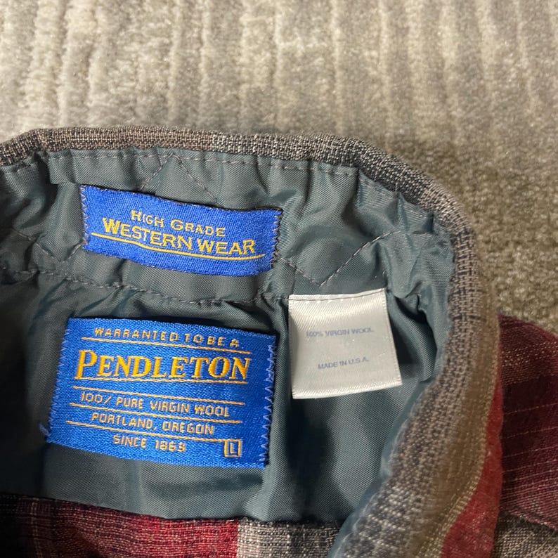 Op de afbeelding: Detail van de binnenkraag van een Pendleton Western Wear shirt. Het shirt heeft een rood en grijs ruitpatroon. Blauwe labels tonen "High Grade Western Wear" en "Pendleton 100% Pure Virgin Wool". Gemaakt in de VS.