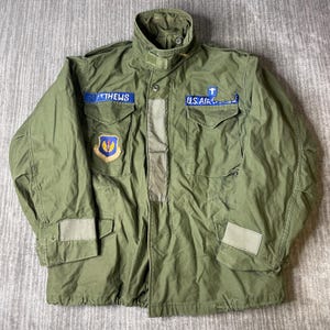 May include: An olive green military-style field jacket featuring a high collar, long sleeves, and four front pockets. The jacket displays a blue name patch reading MATTHEWS, a blue U.S. AIR FORCE patch with a cross symbol, and a colorful shield-shaped patch.