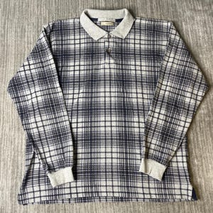 Pode incluir: Uma camisa polo de manga comprida com um padrão xadrez em tons de azul marinho escuro e cinza claro. A peça inclui uma gola cinza claro sólida, punhos canelados e uma carcela de dois botões no decote com botões escuros. O tecido tem uma aparência de malha texturizada em toda a peça.