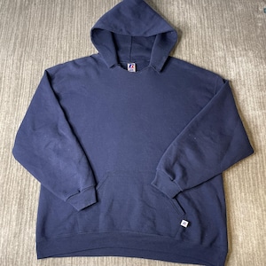 May include: A solid navy blue long-sleeve hooded sweatshirt featuring a front kangaroo pocket, ribbed cuffs, and a ribbed hem. The garment includes a Russell brand tag at the inner neckline and a small white brand logo tab on the front pocket.